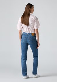 Indigo blue denim jeans with a slim fit, five pockets, and a tan leather patch at the back waistband, paired with a pink striped shirt.