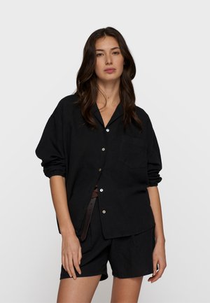 Kobieta o długich ciemnych włosach, ubrana w luźną czarną koszulę na guziki z podwiniętymi rękawami oraz dopasowane czarne szorty, na jednolitym białym tle.