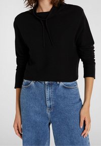 Svart croppad sweatshirt med dragsko i huvan, tillsammans med högmidjade blå denimjeans. Mjuk textur, avslappnad passform och långa ärmar.