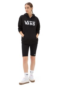 Felpa nera con cappuccio e logo Vans bianco, abbinata a pantaloni biker neri. Gli accessori includono calzini bianchi e sneakers con suola beige.