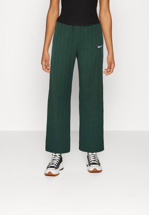 Personne portant un pantalon large côtelé vert foncé, un débardeur noir et des baskets noires et blanches, debout devant un fond uni.