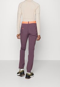 Pantalones outdoor morados con una cinturilla naranja, puños con cordón y bolsillos traseros con cremallera. Combinados con una camiseta de manga larga beige texturizada y zapatillas.