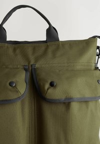 Platnena torba olivno zelene barve z dvema sprednjima žepoma, zaprtima s črnimi zaponkami, in ojačanimi sivimi robovi. Ima dvojne zgornje ročaje.