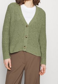 Grüner gestrickter Cardigan mit tiefem V-Ausschnitt, fünf braunen Knöpfen, gerippten Bündchen und Saum sowie einer lässigen Passform, kombiniert mit braunen Hosen.