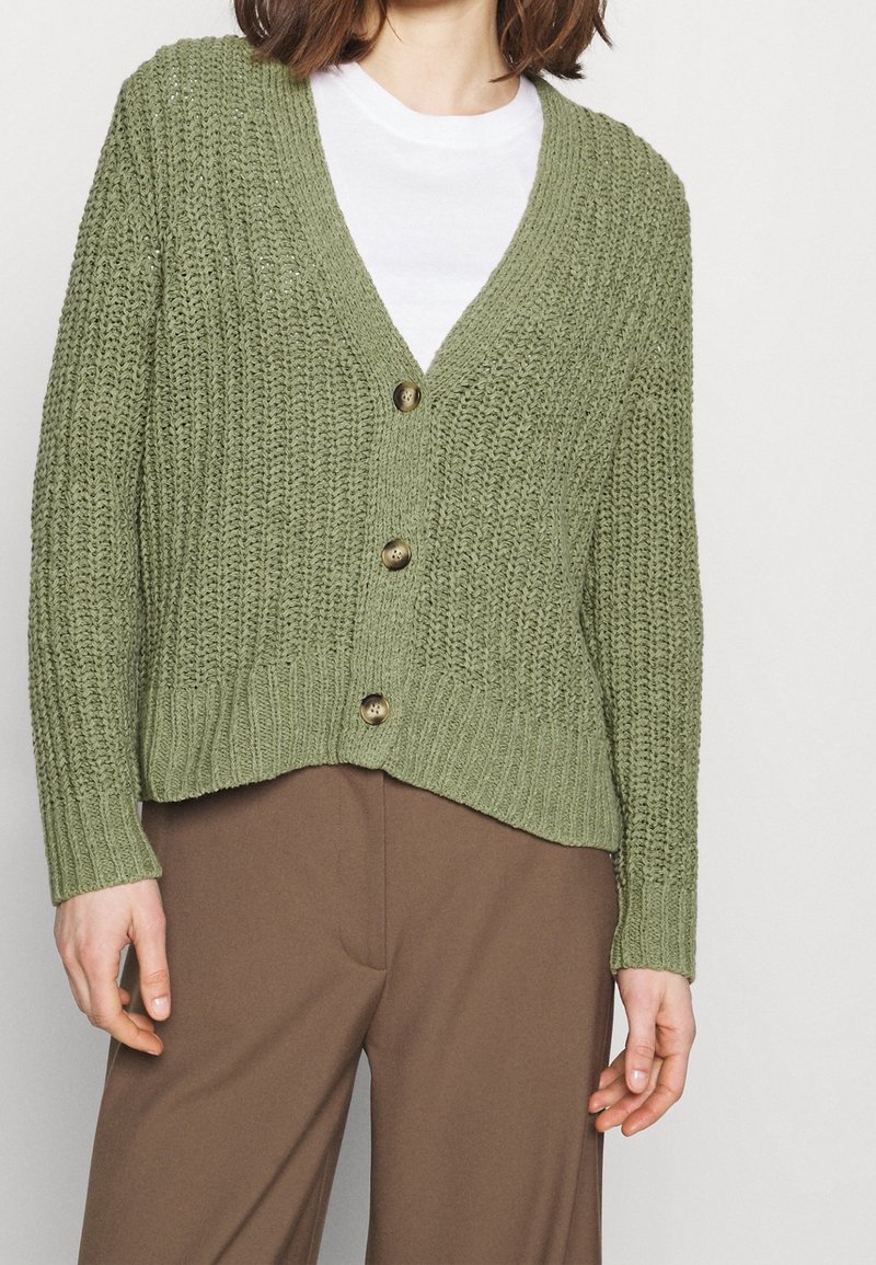 Grüner gestrickter Cardigan mit tiefem V-Ausschnitt, fünf braunen Knöpfen, gerippten Bündchen und Saum sowie einer lässigen Passform, kombiniert mit braunen Hosen.