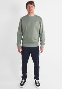 Sweatshirt en coton vert olive avec col rond, poignets et ourlet côtelés. Porté avec un pantalon bleu marine foncé et des baskets noires avec des semelles blanches.