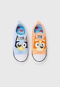 Des chaussures slip-on bleu clair et orange en tissu pelucheux, avec des visages de chiens de dessin animé. Semelles blanches en caoutchouc avec des accents noirs et un étiquetage.