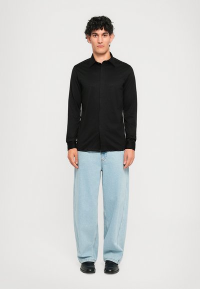 Ung mann med krøllete hår iført en svart langermet skjorte med knapper og lyseblå vide jeans, stående mot en ensfarget bakgrunn.