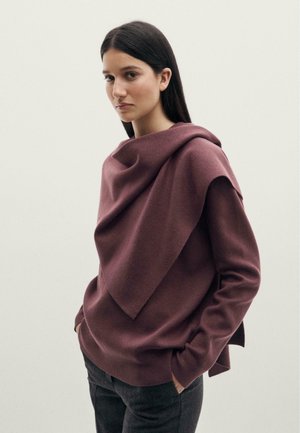 Femme aux longs cheveux foncés portant un pull cache-cœur mauve et un pantalon sombre, debout avec les mains partiellement dans les poches sur un fond clair.