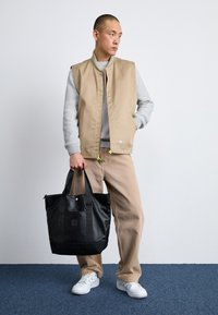 Gilet rembourré beige sur un pull gris, pantalon marron clair et baskets blanches. Tenant un grand sac fourre-tout noir avec un logo. Fond uni.