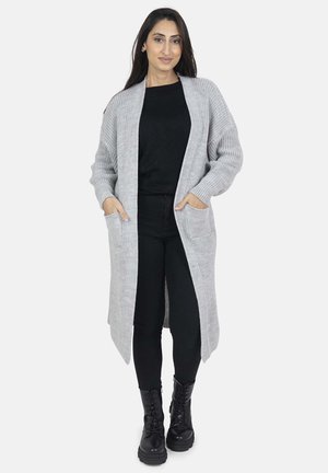Cardigan grigio lavorato a maglia con silhouette lunga, apertura frontale, due tasche laterali e texture a coste. Indossato sopra un top nero e pantaloni neri.