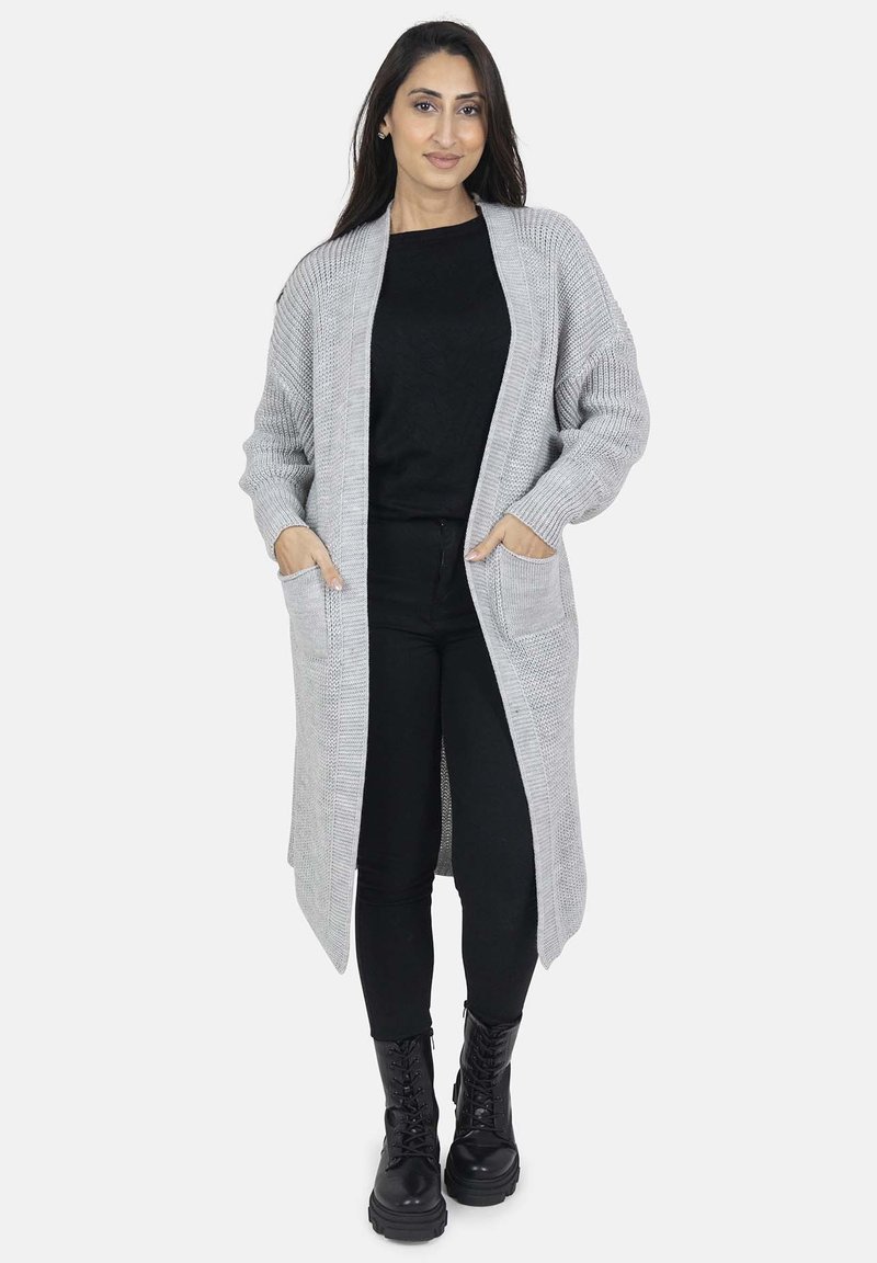 Cardigan grigio lavorato a maglia con silhouette lunga, apertura frontale, due tasche laterali e texture a coste. Indossato sopra un top nero e pantaloni neri.