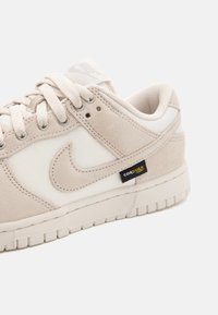 Ténis Nike bege com uma mistura de materiais em camurça e malha, uma entressola branca e um rótulo preto Cordura na lateral.