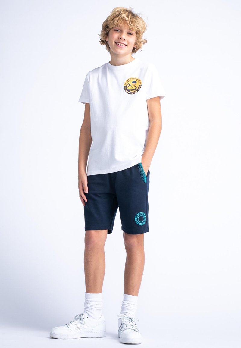 Biała bawełniana koszulka z okrągłym żółtym logo na klatce piersiowej, zestawiona z granatowymi szortami z turkusowymi akcentami i bocznymi kieszeniami.