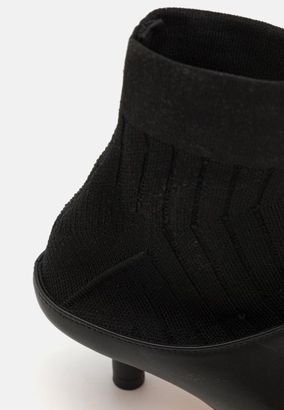 Melnas potītes kurpes ar adītu tekstūru augšdaļā, gludu sintētisku zoli un mazu, noapaļotu papēdi. Iezīmējas ar ciešu piegulšanu un vertikālām ribām.