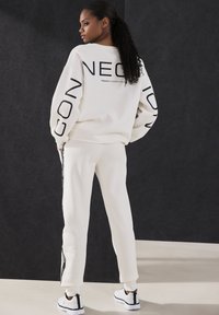 Weißer Baumwoll-Sweatshirt mit schwarzer Beschriftung auf den Ärmeln, kombiniert mit der passenden Hose und Sneakers mit schwarzen Akzenten. Glatte Textur, lässige Passform.