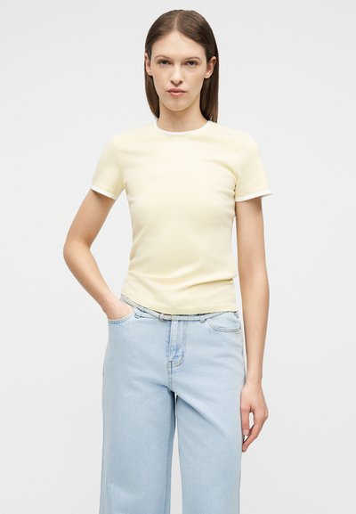 Γυναίκα με ίσια καστανά μαλλιά φοράει κοντομάνικη ανοιχτόκκινη μπλούζα, μπουσουρισμένη μέσα σε ανοιχτόχρωμο μπλε τζιν παντελόνι με ψηλή μέση και φαρδιά μπατζάκια, συνοδευόμενο από λεπτή ζώνη.