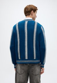 Μπλε ζα κέτα από χνουδωτό ύφασμα με κάθετες λευκές ρίγες, πλεκτή υφή και ριμπ γιακά. Φορεμένη πάνω από γκρι τζιν, δείχνοντας ένα άνετο, χαλαρό στυλ.