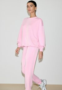 Hele roosa suurendatud suurusega pikkade varrukatega dressipluus, sobitatud samast pehmest kangast pükstega, millel on lõdvestunud lõige ja elastsed mansetid.