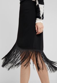 Černá sukně vyrobená z hladkého materiálu, s třásněmi podél lemu a elegantním střihem, který končí nad koleny.