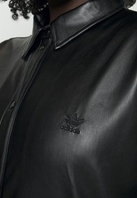 Černá kožená košile s designem na zapínání na knoflíky a s špičatým límcem. Na hrudi se nachází razítkované logo Adidas. Hladká textura.