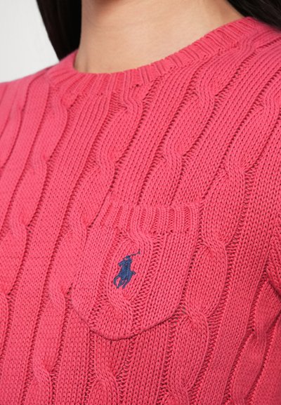Ροζ πουλόβερ με πλέξη καλώδιο, στρογγυλή λαιμόκοψη και μπροστινή τσέπη. Περιλαμβάνει μικρό μπλε σκούρο λογότυπο στην τσέπη, που αναδεικνύει το υφασμάτινο ανάγλυφο μοτίβο.