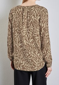 Femme portant un chemisier beige à manches longues avec un motif abstrait en zigzag marron et un pantalon noir, debout contre un mur clair texturé.