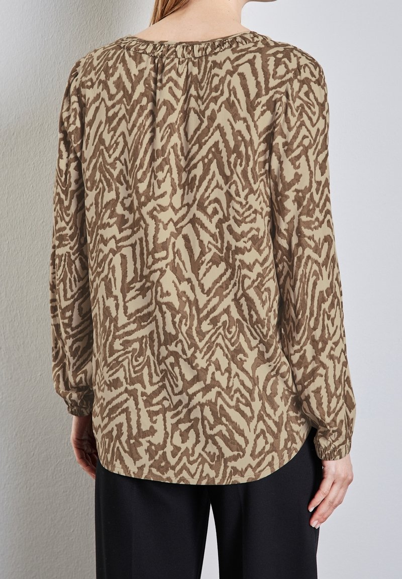 Femme portant un chemisier beige à manches longues avec un motif abstrait en zigzag marron et un pantalon noir, debout contre un mur clair texturé.