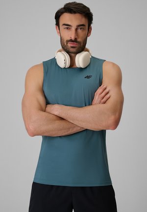 Atletyczny mężczyzna z brodą w niebieskiej koszulce bez rękawów i czarnych szortach, białe słuchawki na szyi, skrzyżowane ramiona, neutralne szare tło.