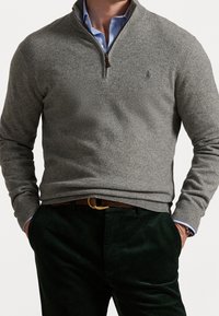 Pull-over en polaire grise avec col demi-zip, poignets et ourlet côtelés, associé à un pantalon en velours côtelé vert foncé. Présente une broderie de logo sur la poitrine.