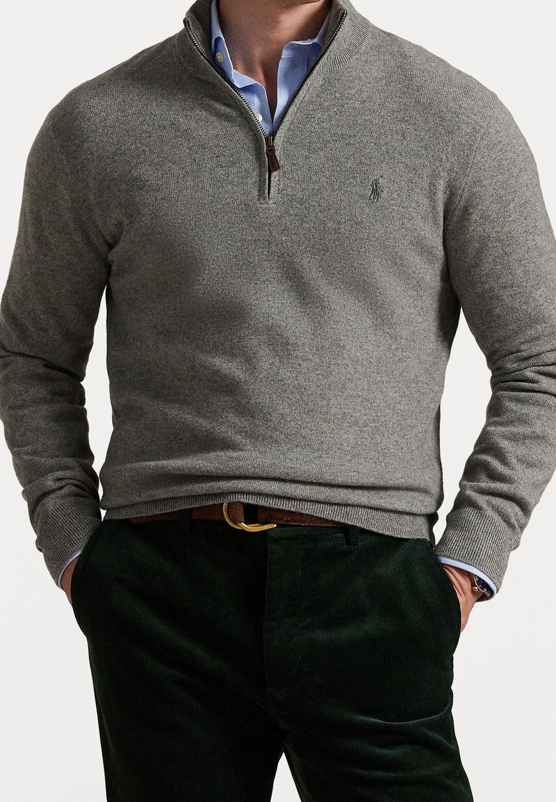 Pull-over en polaire grise avec col demi-zip, poignets et ourlet côtelés, associé à un pantalon en velours côtelé vert foncé. Présente une broderie de logo sur la poitrine.