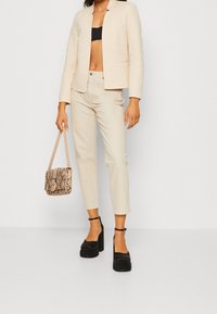 Blazer ajustado beige sobre un top corto negro, combinado con pantalones crema y tacones de plataforma negros. El bolso tiene un diseño de estampado de serpiente.