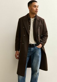 Abrigo marrón de doble botonadura con un acabado texturizado, llevado sobre una camisa crema y vaqueros azules, con solapa de pico y bolsillos frontales.