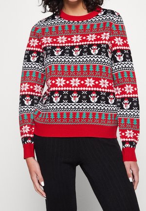 Femme portant un pull de Noël rouge avec des flocons de neige, des bonhommes de neige et des motifs d’arbres verts, associé à un pantalon noir.