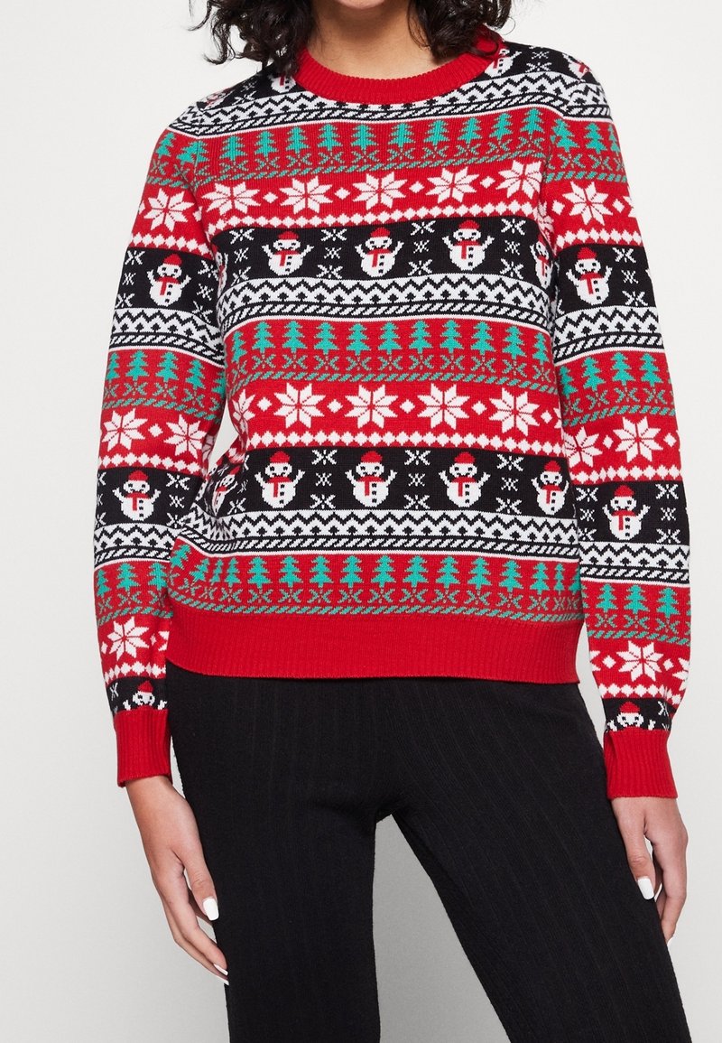 Femme portant un pull de Noël rouge avec des flocons de neige, des bonhommes de neige et des motifs d’arbres verts, associé à un pantalon noir.