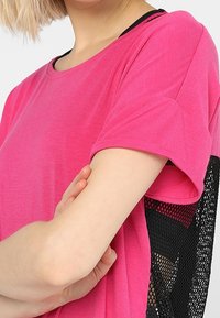 Žena v jasně růžovém tričku s krátkým rukávem a černou síťovinou na straně, se zkříženýma rukama, světlý tón pleti, blond vlasy.