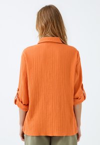 Texturiertes oranges Halsbandhemd mit aufgerollten Ärmeln. Das weiche Material weist senkrechte Linien auf. Das Hemd wird von hinten gegen einen schlichten Hintergrund gezeigt.