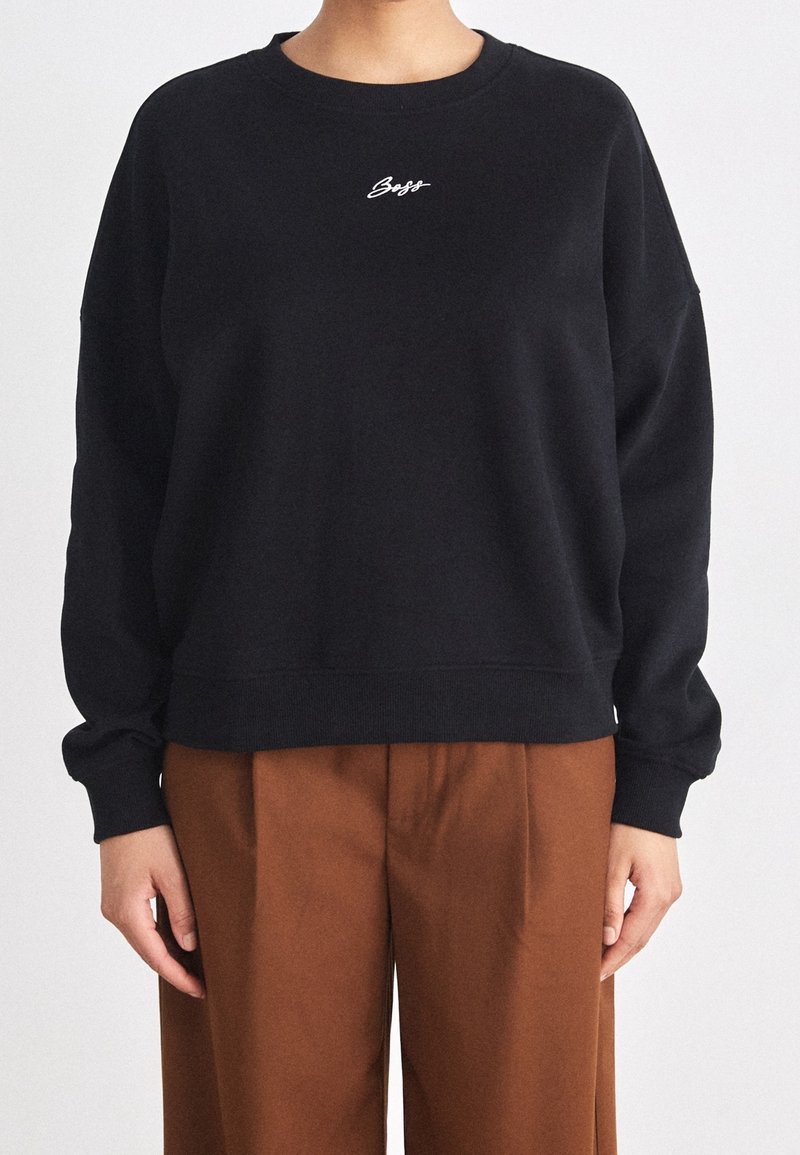 Personne portant un sweat-shirt noir à manches longues avec le texte blanc "Boss" sur la poitrine et un pantalon plissé marron taille haute, debout sur un fond blanc.