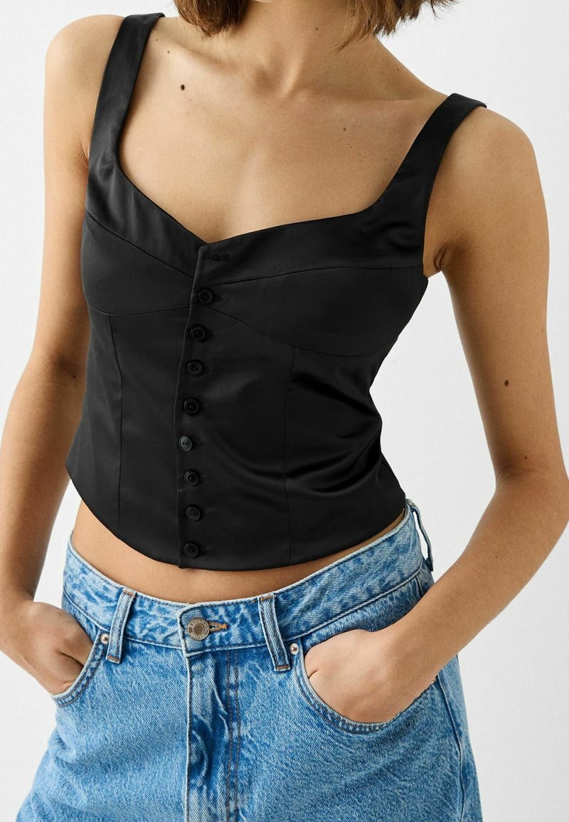 Czarny atłasowy gorset z szerokimi ramiączkami, kwadratowym dekoltem i rzędem sześciu guzików z przodu. Zestawiony z wysokim stanem krótkich spodenek jeansowych.