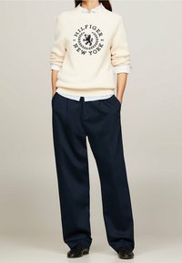 Cream ribbed sweater with navy logo and year "1985," paired with navy wide-leg trousers. Layered over a light blue collared shirt.