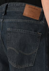 Jeans de denim oscuro con una etiqueta de marca de cuero en la cintura trasera, que presentan diseños de bolsillo bordados y un estilo clásico de cinco bolsillos.