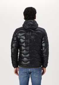 Giacca a vento nera con design trapuntato e cappuccio, caratterizzata da una texture lucida e polsini elastici. Indossata con jeans blu.