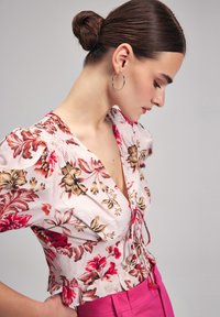 Bluse mit floralem Muster in hellrosa, mit kurzen Puffärmel, V-Ausschnitt und gerafftem Oberteil. Kombiniert mit hoch taillierten rosa Hosen.