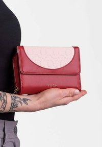 Red and pink leather wallet with rounded flap, zipper pocket, and embossed pattern. Gold "VUCH" logo on front. Hand-held for scale.