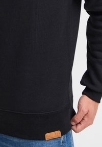 Sudadera negra con puños y dobladillo acanalados. Presenta una etiqueta de cuero marrón en la parte inferior. La tela parece suave y lisa.