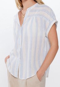 Chemise en lin à rayures verticales bleu clair et blanches, avec fermeture boutonnée sur le devant, design sans col et manches courtes retroussées.