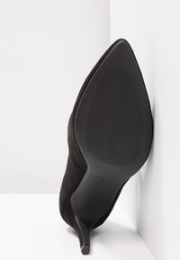 Chaussure à talons hauts en suède noir avec un bout pointu et un fin talon aiguille. La semelle est texturée avec un motif ondulé, affichant le logo de la marque.