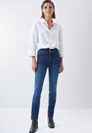 Jean en denim bleu foncé avec une coupe slim, doté de cinq poches et d'une fermeture à bouton. Associé à une chemise blanche à manches longues ample.