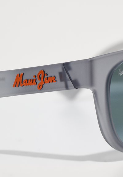 Pelēkas plastmasas saulesbrilles ar nedaudz caurspīdīgu rāmi. Rokas daļā ir oranžs "Maui Jim" logo. Objektīvi ir tumši zaļi.