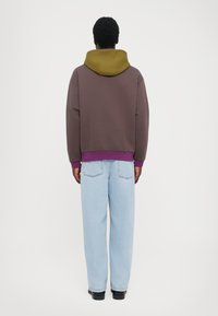 Felpa color block marrone e oliva con accenti viola, vestibilità rilassata, polsini a costine e jeans azzurri a gamba dritta. Sfondo semplice.